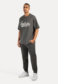 Grå oversized t-shirt med hvid "Smilodox" tekst, parret med matchende tapered sweatpants. Fodtøj inkluderer hvide sneakers. Enkel design.
