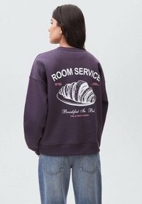 Kvinna med långt mörkt hår iklädd en lila sweatshirt med "ROOM SERVICE" och en grafik av en croissant på ryggen, tillsammans med blå jeans.