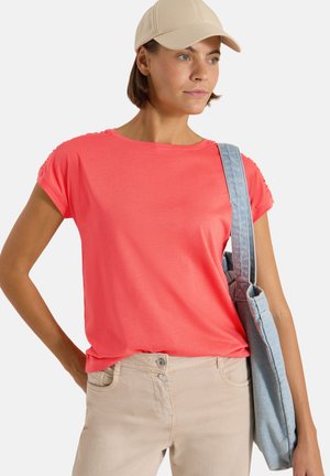 Femme portant une casquette beige, une chemise corail à manches courtes, un pantalon beige clair, portant un sac cabas en denim bleu clair sur l'épaule, regardant sur le côté.