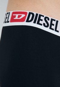 Boxer-briefs en coton noir avec une taille élastique blanche arborant le logo "DIESEL" en noir et un accent rouge. Texture lisse.