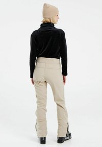 Top lungo in velluto nero con collo alto, abbinato a pantaloni beige a vita alta con zip laterali e stivali da sci neri.