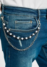 Cadena de plata sujeta a unos vaqueros, con cuentas en blanco y negro, algunas que parecen dados. Denim desgastado con un acabado azul y cierre de botón.