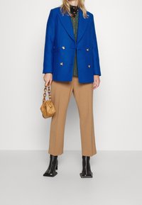 Blazer blu doppiopetto, maglione verde testurizzato, pantaloni beige, stivali neri con tacco a blocco e una piccola borsa marrone con manico a catena.