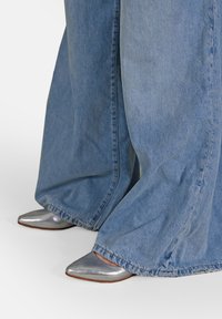 I pantaloni in denim a gamba larga si incontrano con scarpe a punta in metallo argentato, dalla texture liscia e suola piatta, creando un effetto visivo di contrasto.