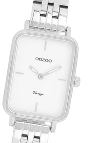 Montre rectangulaire en métal argenté avec un cadran blanc, comportant des repères horaires minimalistes, des aiguilles, et le texte "OOZOO Vintage". Bracelet avec texture.