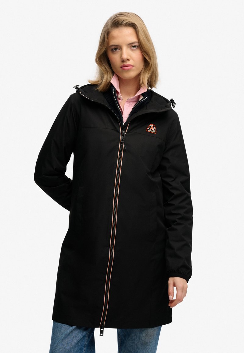 Abrigo impermeable negro con cierre de cremallera, capucha y logo naranja. Presenta puños elásticos y un diseño simple y recto.