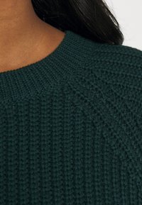 Grön stickad tröja med ett strukturerat mönster, ribbad halsringning och raglanärmar, som framhäver tygets tjocklek och värme.