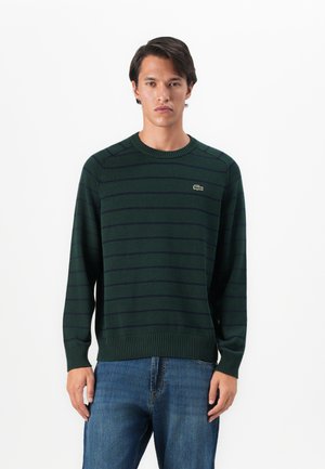 Mørkegrønn stripete genser med ribbet hals og mansjetter; har en liten logo på venstre bryst. Kombinert med blå denimjeans.