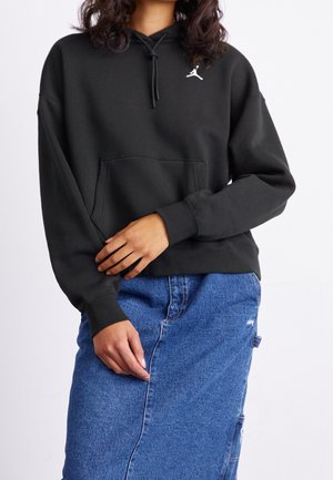 Personne portant un sweat à capuche noir avec un logo blanc et une jupe en jean bleue, debout devant un fond clair uni.