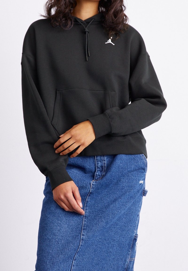 Personne portant un sweat à capuche noir avec un logo blanc et une jupe en jean bleue, debout devant un fond clair uni.