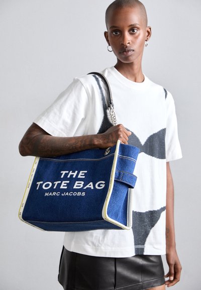 Denima iepirkumu soma ar baltu uzrakstu "THE TOTE BAG MARC JACOBS", ar kontrastējošu dzeltenu apdari un izturīgu melnu rokturi.
