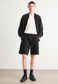 Schwarze Bomberjacke mit Reißverschluss, weißes T-Shirt, schwarze strukturierten Shorts, schwarze Socken mit rotem Schriftzug und schwarze Sneakers. Einfaches, lässiges Outfit.