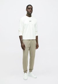 Balta, garām piedurknēm krekls ar logotipu uz krūtīm, kopā ar bēšām, šaurām biksēm un baltām sporta kurpēm. Vienkāršs fons.