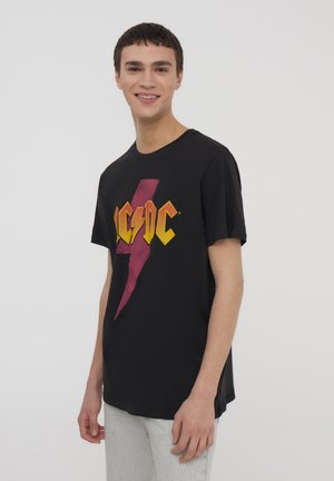 T-shirt in cotone nero con un audace logo AC/DC giallo e rosso con design a fulmine. Maniche corte, collo tondo, vestibilità rilassata.