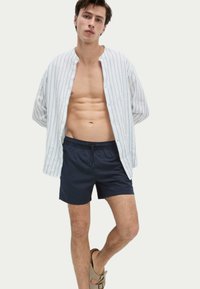 Camisa blanca con rayas azules, desabotonada, llevada sobre pantalones cortos de baño azul marino. El modelo está descalzo, con sandalias marrones. Diseño sencillo y casual.