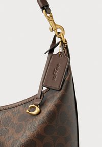 Sac à main en cuir marron arborant un motif Coach, une finition en métal doré et une étiquette amovible attachée par une chaîne. Forme incurvée avec une surface texturée.