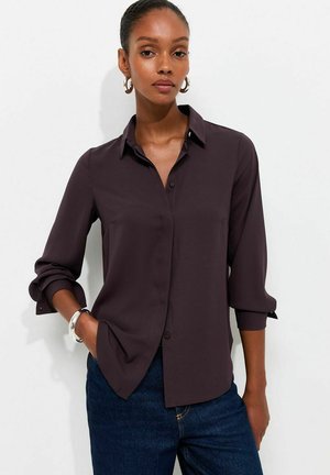 Femme portant une chemise boutonnée à manches longues sombre avec manches retroussées, un jean bleu, des boucles d'oreilles anneaux et un bracelet argenté sur un fond uni.