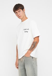 Witte katoenen t-shirt met korte mouwen en geborduurde tekst "rock me, Baby" op de linkerkant van de borst; gecombineerd met lichte spijkerbroek.