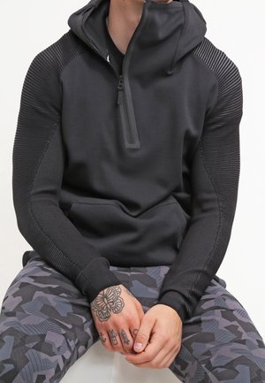 Homme portant un sweat à capuche noir à demi-fermeture éclair avec des manches texturées et un pantalon à motif camouflage, les mains reposant sur les genoux montrant des doigts tatoués.