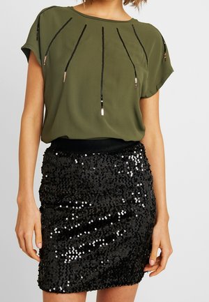 Mujer con top de manga corta verde oliva con líneas verticales de lentejuelas negras y minifalda negra con lentejuelas sobre un fondo liso.