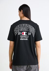Melns kokvilnas t-krekls ar boldero pelēko uzrakstu "CHAMPION", gada "1919" un logotipu sarkanas un baltas krāsas. Brīvs griezums. Īsas piedurknes.