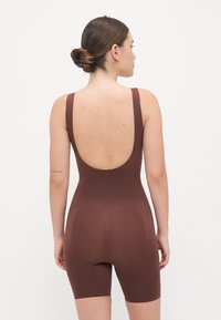 Rudas bodysuit'as su žemu nugara ir plačiomis petnešėlėmis, pagamintas iš lygaus, tamprių medžiagų. Turi prigludusį dizainą ir vidutinio ilgio iki šlaunų.