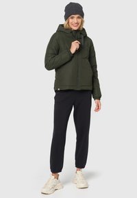 Groene gewatteerde jas met een capuchon, voorkant rits en elastische manchetten. Gecombineerd met zwarte joggers en lichtgekleurde sneakers. Gebreide beanie op het hoofd.