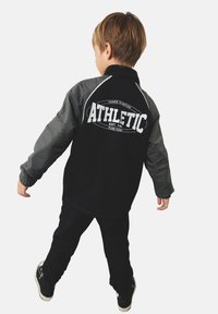 Zwart en grijs sportjack met lange mouwen, voorzien van witte tekst "ATHLETIC" op de rug en contrasterende schouderaccenten. Zwarte broek en sneakers.