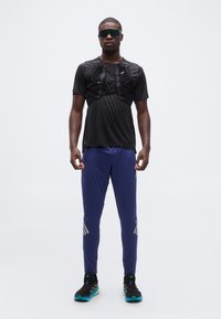 Άνδρας που φοράει μαύρο αθλητικό τοπ με ιμάντα, μπλε παντελόνι φόρμας, μαύρα αθλητικά παπούτσια και μεγάλα αντανακλαστικά γυαλιά ηλίου αθλητικού τύπου, στέκεται μπροστά σε λευκό φόντο.