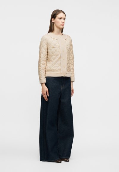 Γυναίκα με μακριά καστανά μαλλιά, που φοράει έν α μπεζ μπουφάν με κεντήματα και σκούρο τζιν με φαρδιά μπατζάκια, στέκεται μπροστά σε έναν απλό φωτεινό φόντο.