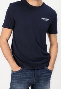 Man wearing navy blue short-sleeve T-shirt with small white logo on chest and blue denim jeans, hands tucked into pockets.