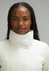 Biała kurtka puchowa z wysokim kołnierzem, gładką teksturą i minimalnymi przeszyciami. Noszona przez osobę z warkoczami.