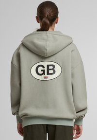 Oliwkowy, oversize'owy hoodie z czarnym owalnym logo z napisem "GB" i małą flagą Union Jack. Posiada kieszeń kangurka i ściągacze na rękawach.