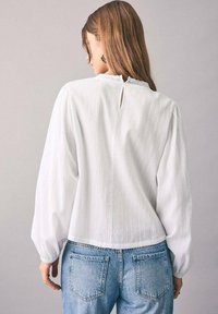 Blusa bianca con maniche lunghe in tessuto texturizzato, scollatura rotonda e dettaglio con apertura a chiave sul retro, abbinata a jeans di denim azzurro chiaro.