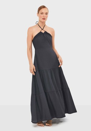 Mujer vestida con un vestido largo negro sin mangas con cuello halter y falda de volantes, de pie descalza contra un fondo claro y liso.