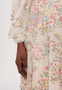 Robe en broderie en tissu transparent avec des motifs floraux en rose, bleu et jaune. Elle présente des manches bouffantes et une finition en dentelle aux poignets.
