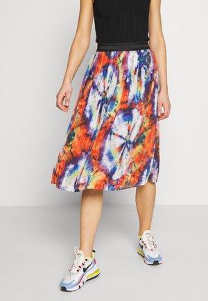 Person iført en farverig plisseret midi nederdel med tie-dye mønster, sort ærmeløs top og hvide Nike sneakers stående på gråt gulv.