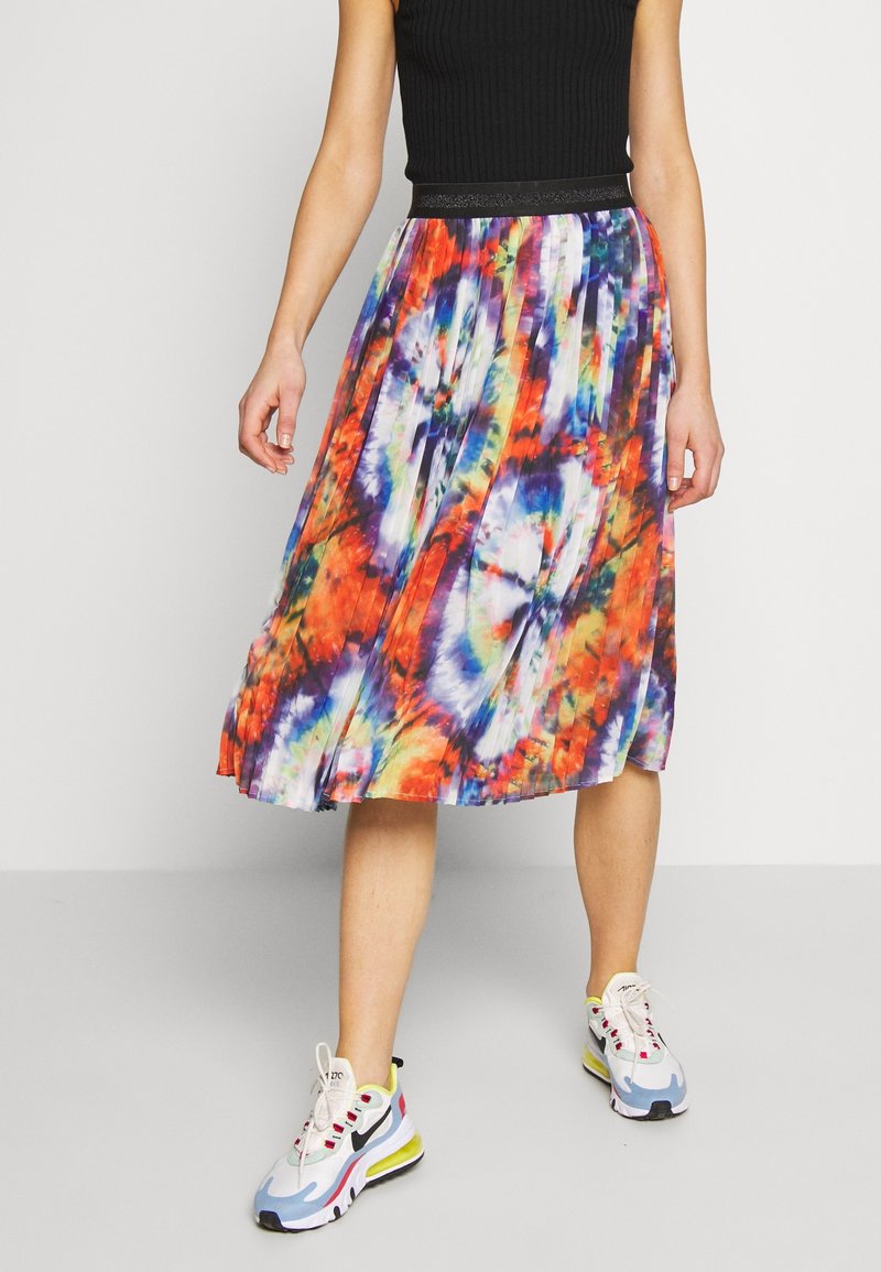 Personne portant une jupe midi plissée colorée avec un motif tie-dye, un haut noir sans manches et des baskets blanches Nike, debout sur un sol gris.