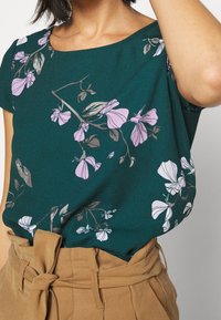 Blusa floreale verde con fiori rosa chiaro e foglie grigie. Tessuto morbido con vestibilità rilassata e scollatura ampia. Abbinata a pantaloni beige con cintura.