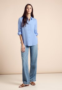 Camisa de botones azul claro, con las mangas remangadas, combinada con unos jeans de mezclilla claros de pierna ancha y sandalias planas marrones sobre un fondo neutro.