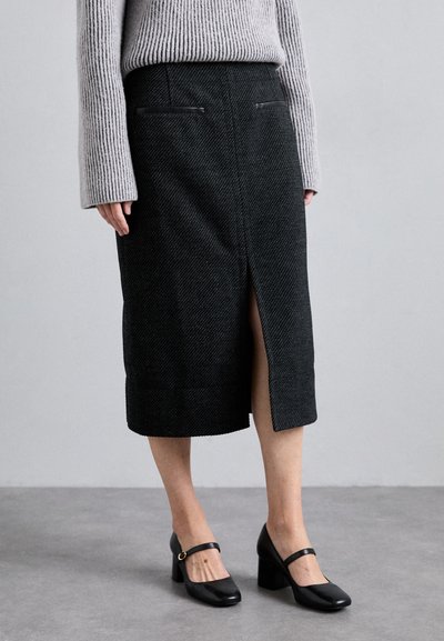 Jupe midi en pied-de-poule noir avec une fente à l'avant et des accents en cuir synthétique sur les poches. Associée à des chaussures à talons noires et un pull gris clair.