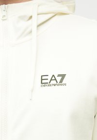 Λευκό φούτερ με φερμουάρ από απαλό ύφασμα, με ανάγλυφο λογότυπο EA7 Emporio Armani σε σκούρο πράσινο. Περιλαμβάνει κορδόνια στο κουκούλα.