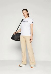 Mladá žena v bielej tričku Champion, béžových nohaviciach a teniskách, drží čiernu športovú tašku cez rameno na pozadí bez vzoru.