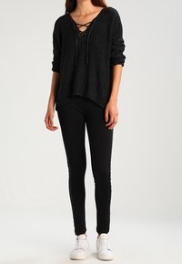 Pull noir en maille avec un décolleté en V et un détail à lacets, associé à un pantalon slim noir et des baskets blanches. Tissu texturé avec une coupe décontractée.