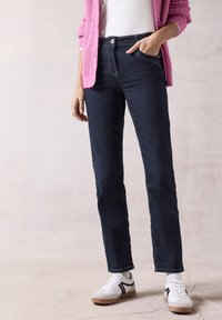 Jeans de mezclilla de un azul oscuro con un diseño de pierna recta, que presentan una cintura alta, cinco bolsillos y costuras amarillas. Combinados con zapatillas blancas.