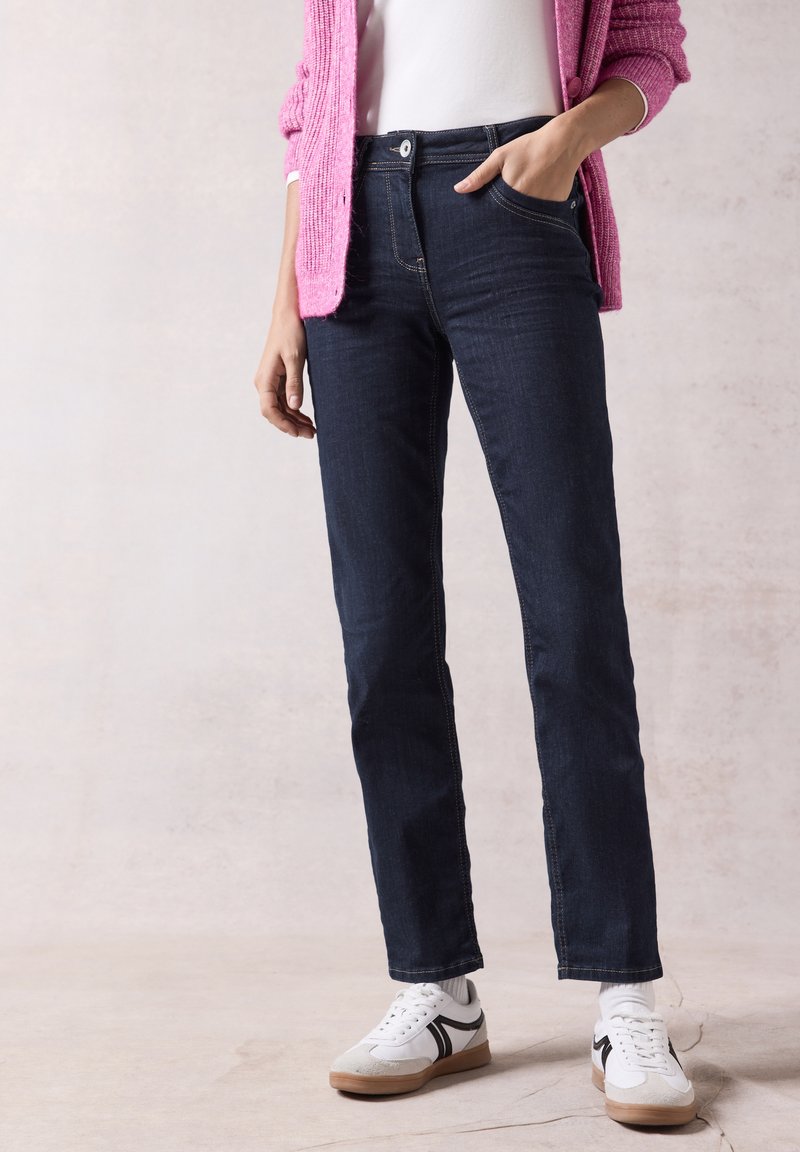 Jeans de mezclilla de un azul oscuro con un diseño de pierna recta, que presentan una cintura alta, cinco bolsillos y costuras amarillas. Combinados con zapatillas blancas.
