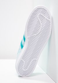 Vitt sneakersula med fiskbensmönster lutad mot en vägg, vilket visar en del av skoens mesh-övre med turkosa ränder.