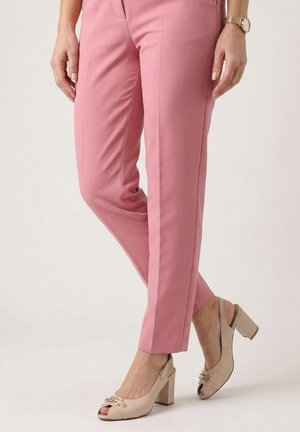 Person iført lyserøde skræddersyede bukser og beige blokhælede sko med åbne tæer, stående med krydsede ben mod en ensfarvet baggrund.