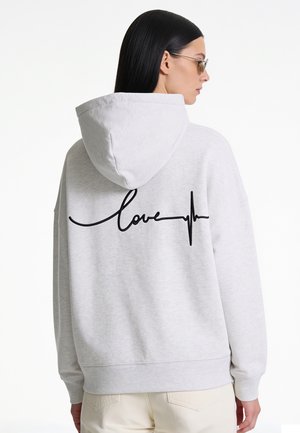 Femme aux cheveux noirs et lisses portant des lunettes de soleil et un sweat à capuche gris clair avec le mot "love" et une ligne de battement de cœur noire dans le dos.