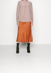 Ljusrosa långärmad blus med volangdetaljer, kombinerad med en silkeslen orange midikjol. Svarta ankelboots fulländar outfiten.